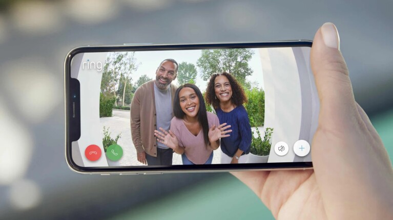 An image of a hand holding a phone that shows three people standing outside a house waving to the Ring doorbell camera. 