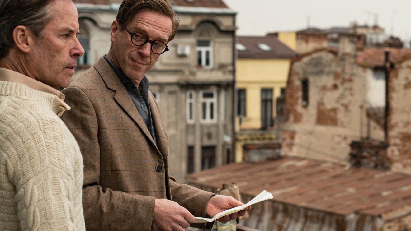 An image of two men standing on a balcony. One is holding a paper and looking at the other man. And the other man is looking off into the distance.