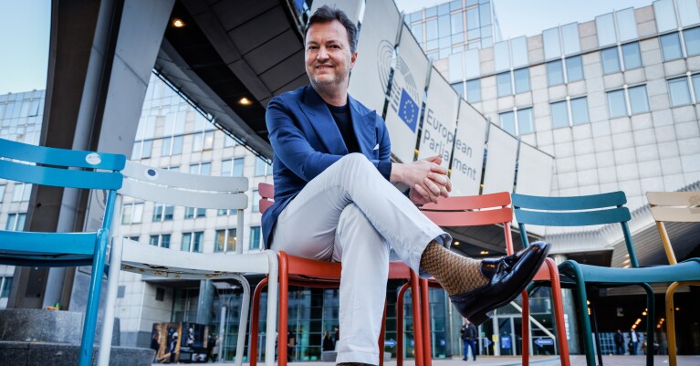 Man in blue blazer sitting on colorful chairs outside European Parliament building