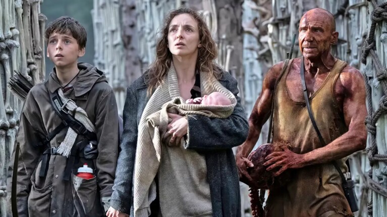 Surrounded by skeletal remains which have been organised into towers, Ralph Fiennes and Alfie Williams stand alongside Jodie Comer, who holds a baby
