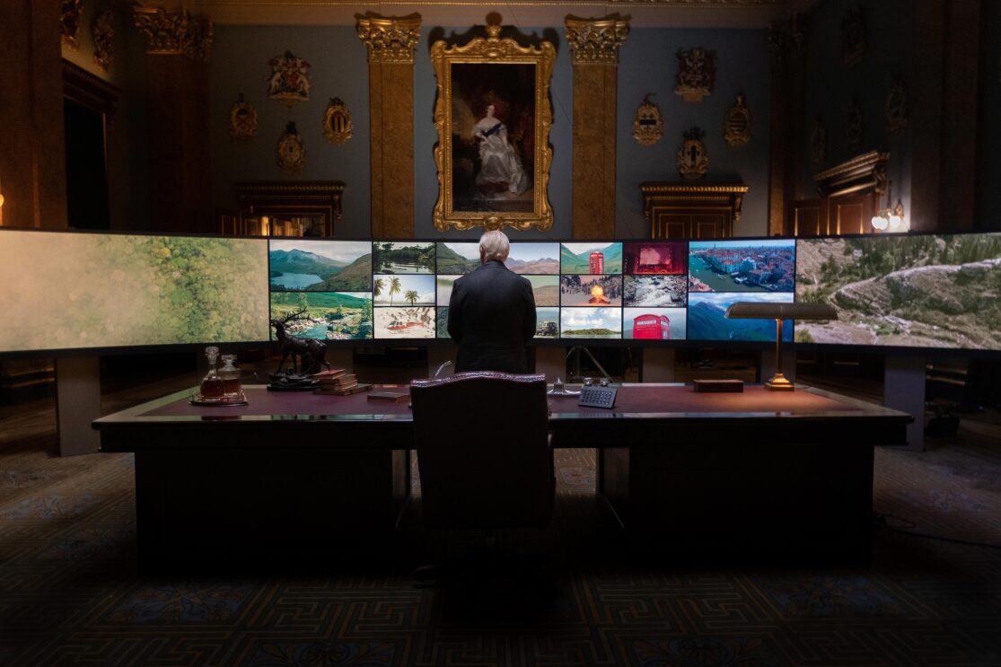 Brian Cox as The Controller stands in front of multiple screens 