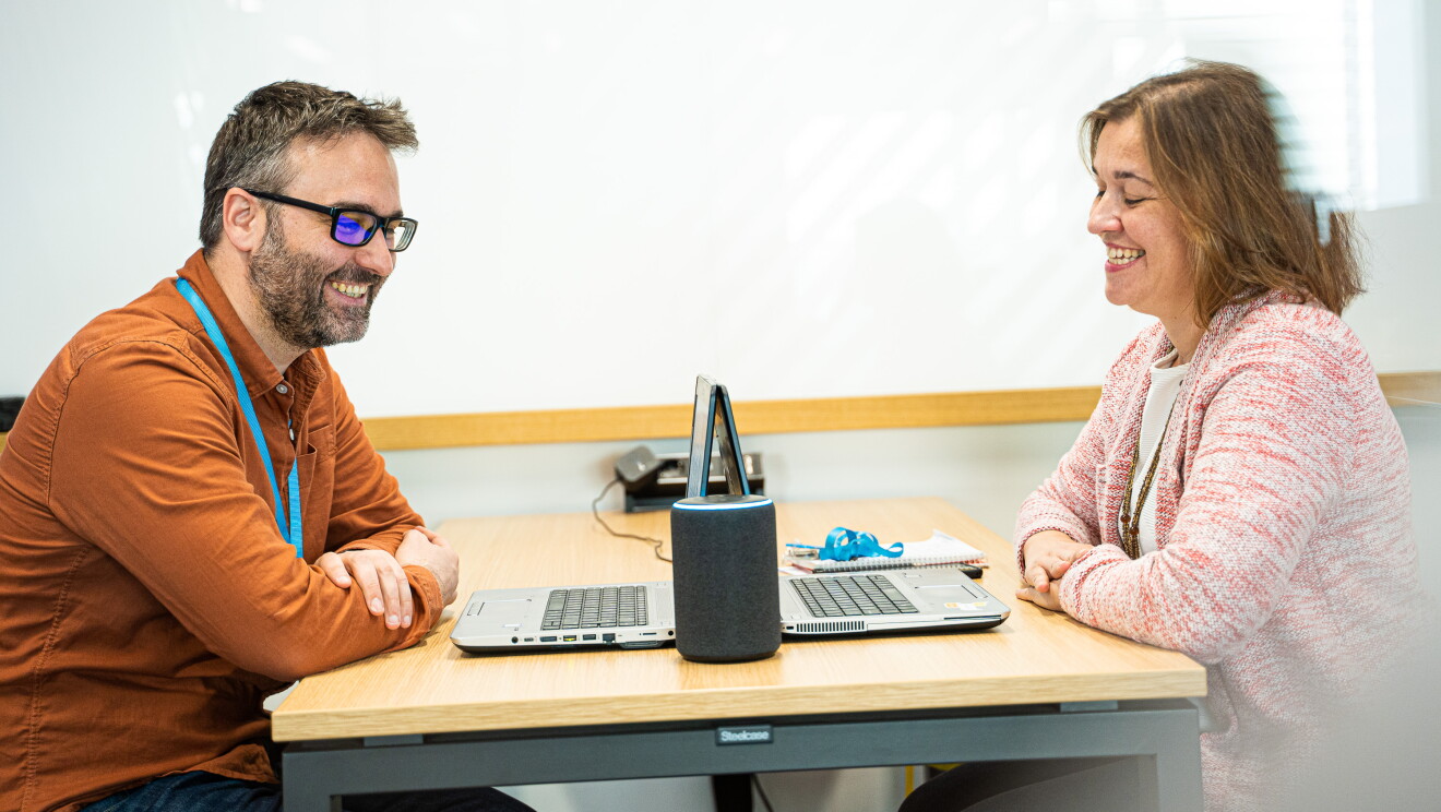 Marta Fernández y Daniel Méndez son Content Program Manager de Alexa. Se encuentran sentados de frende, en una mesa con dos ordenadores y un dispositivo Amazon Echo. Los dos sonrien mirando el dispositivo, tiene los brazos entrecruzados. Daniel con gafas de pasta negra, una camisa marrón y el pelo negro y blanco. Marta con una camiseta blanca y una chaqueta rosa.