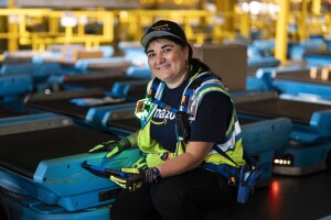 Woman in hat and safety vest posing with blue robotic equipment in Amazon facility.