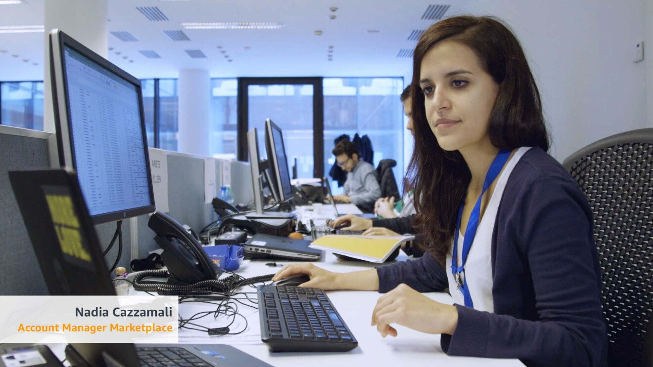 Ritratto di Nadia Cazzamali, Account Manager Marketplace di Amazon. E' seduta alla sua scrivania e guarda a uno dei due schermi che ha di fronte. Sullo sfondo, alla sua destra, due suoi colleghi e le finestre dell'ufficio. 