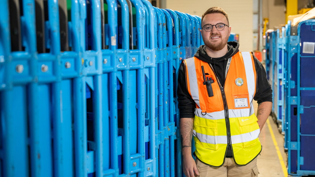 Young warehouse worker in orange and yellow safety vest standing beside blue storage cages