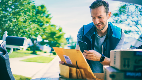 An image of an Amazon delivery driver wearing an Amazon delivery vest and scanning a package using his phone. Behind him is a street view of a neighborhood with green grass and trees.