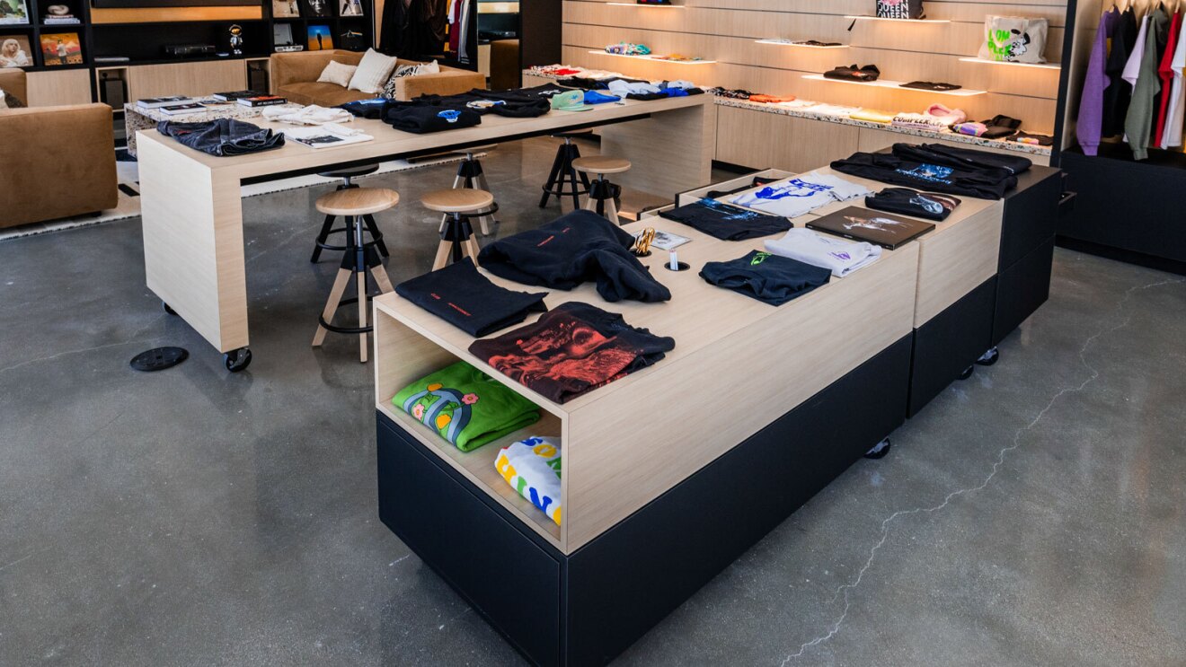 A room with two large tables made of light wood. There are stools set up at one of the tables, and the other table has compartments for sketching tools like pencils and scissors. There are also band t-shirts folded and sitting atop both tables. 