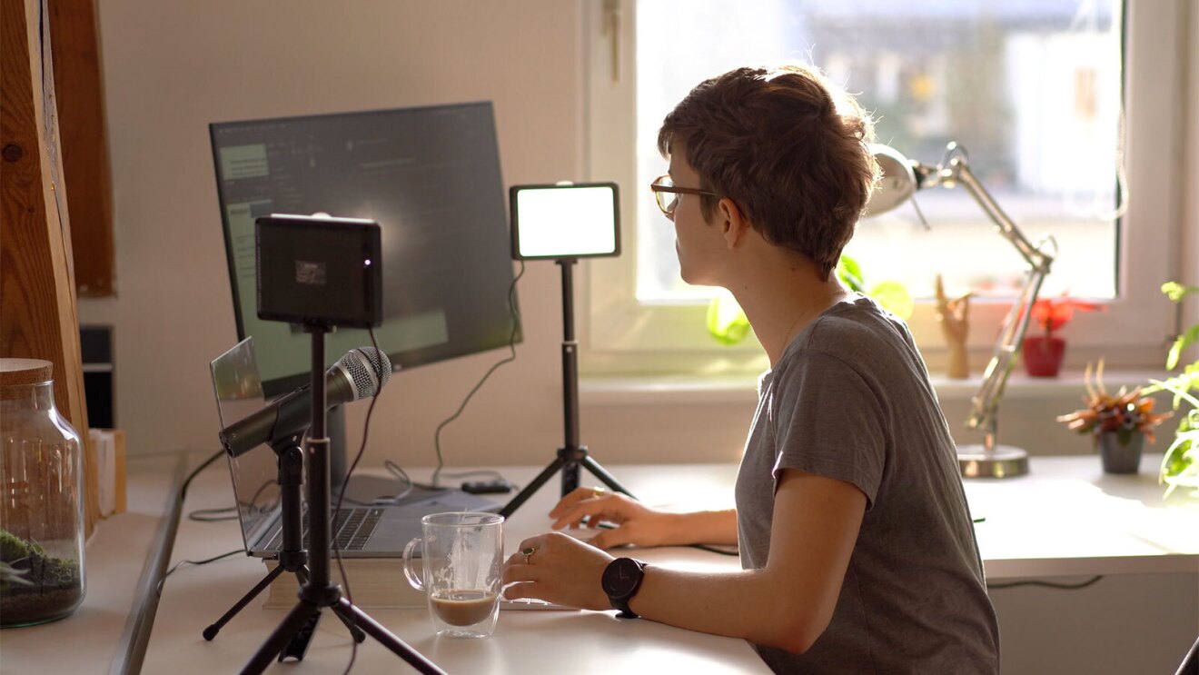 A professional photo of Antonia sitting at her desk with recording equipment surrounding her and an iced coffee in front of her.