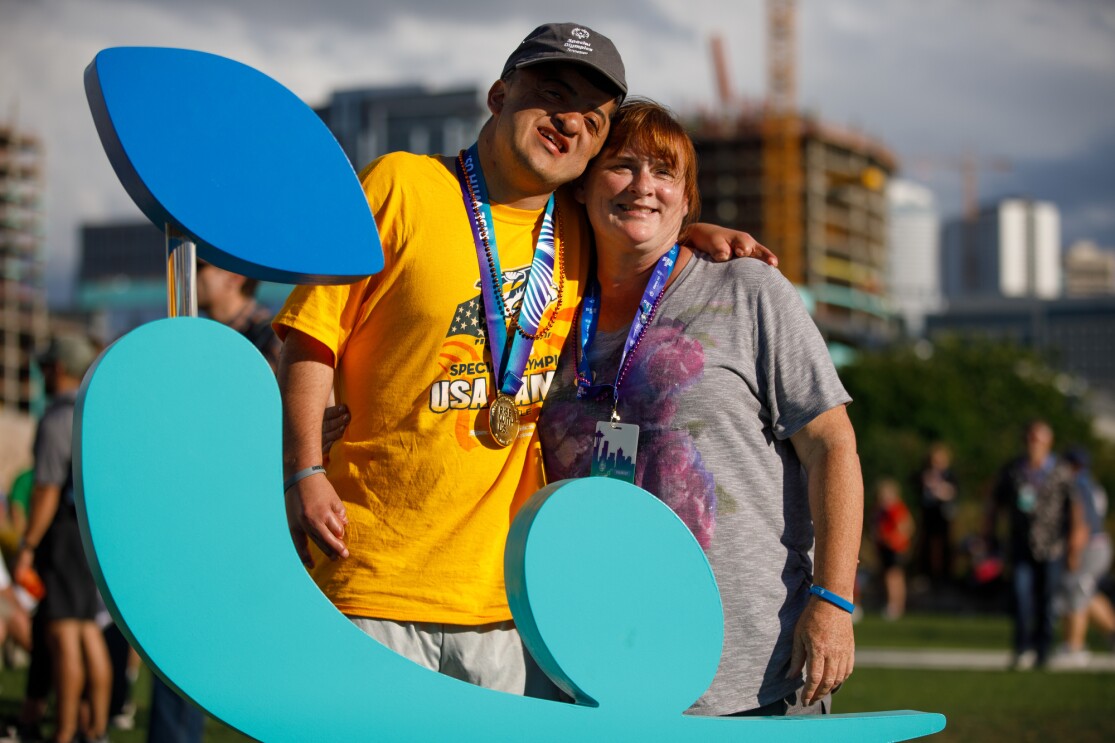A Special Olympics athlete (male) poses for a photo with a female, behind the USA Games logo. Part of the Seattle skyline is visible in the background.