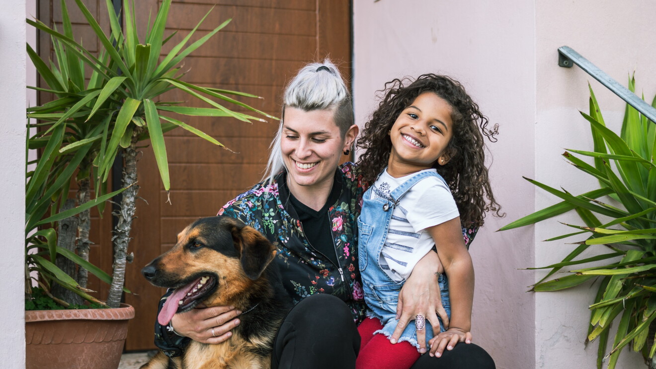 Elisabetta è seduta sugli scalini dell'ingresso della sua abitazione e abbraccia sua figlia, una bimba con i capelli ricci e neri che indossa dei leggins rossi, e il suo cane, un pastore tedesco. Attorno a loro si intravedono delle piante in quello che è il loro giardino. 