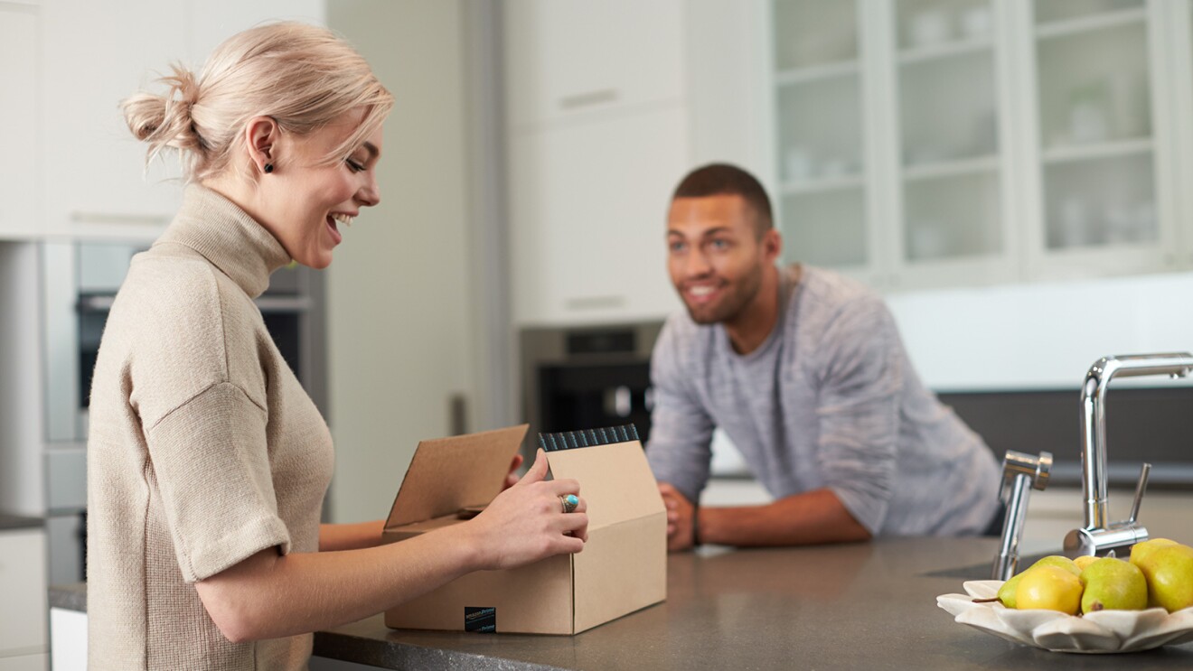 An image of a woman and a man in a kitchen. The woman is opening an Amazon package. 