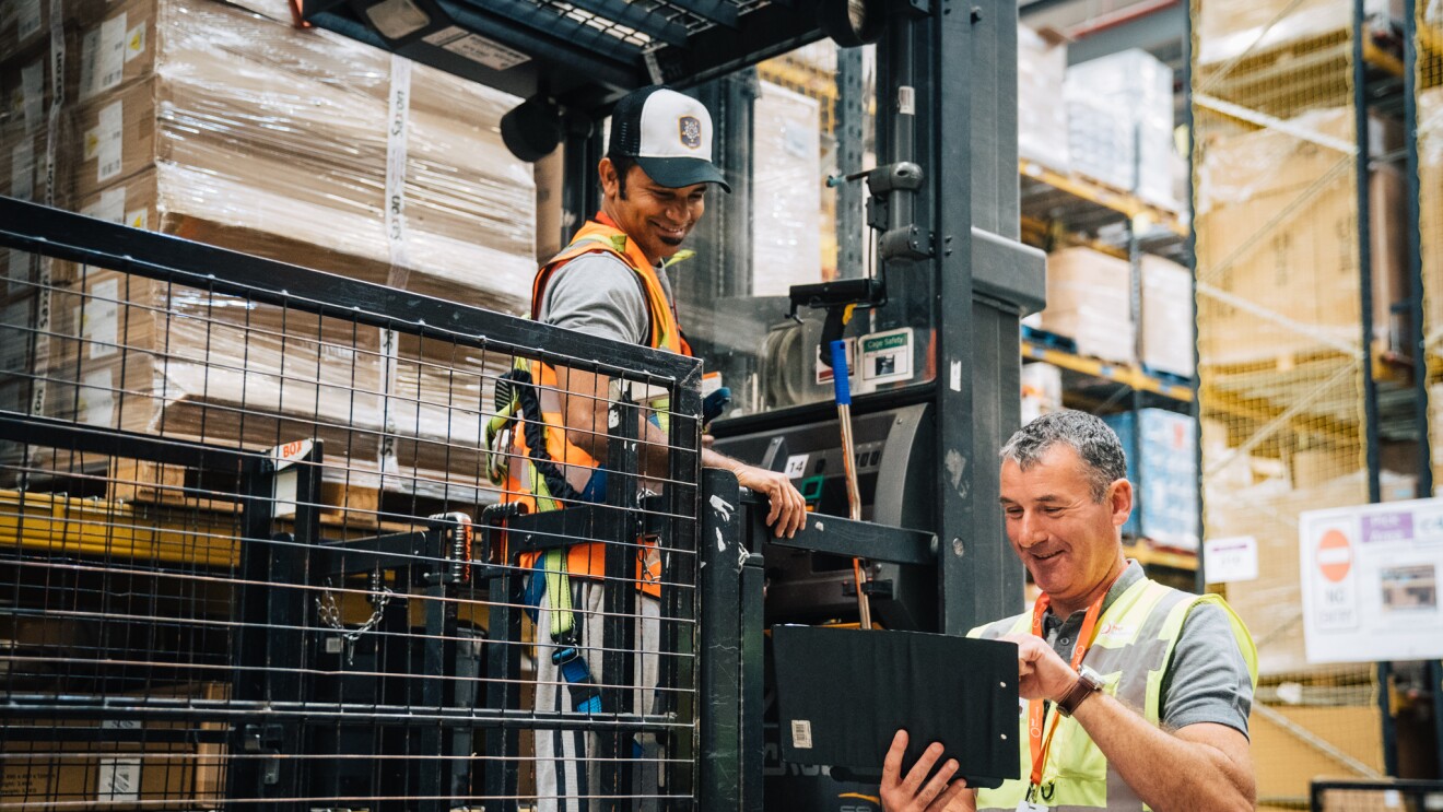 Temporary associate John Sheerin smiling at a colleague in an Amazon Fulfilment Centre and typing on a tablet.