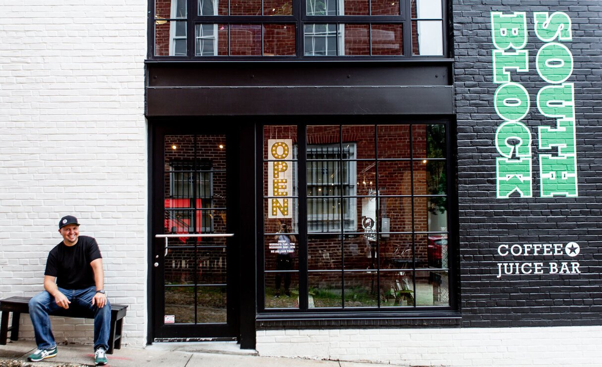 An image of a man smiling for a photo while sitting on a bench outside of a storefront. On the wall of the storefront there is text that reads "South Block Coffee Juice Bar."