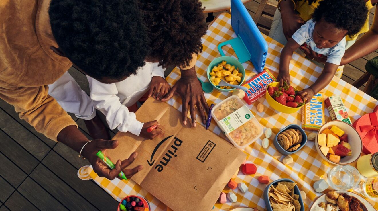 A family sits at a table filled with food and a parent and child draw on a Prime brown grocery bag.