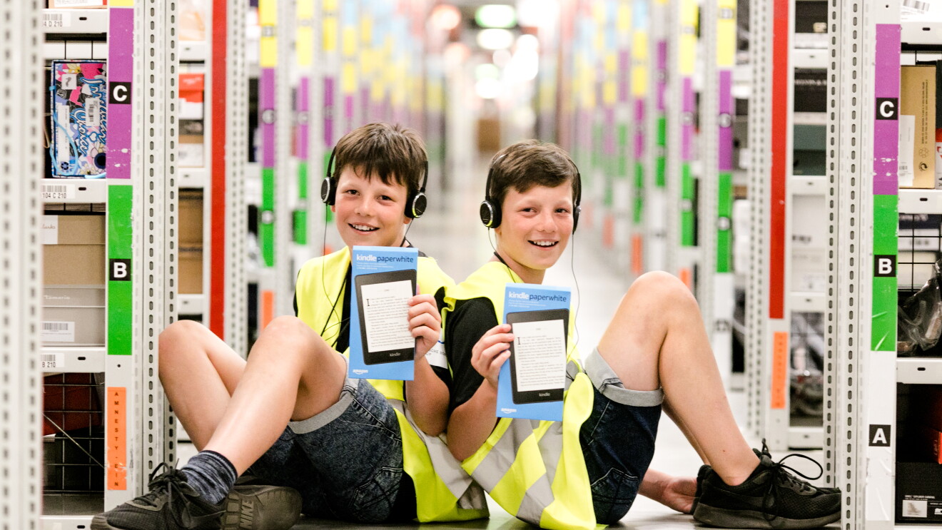 Zwei gleich aussehnde Jungen sitzen Rücken an Rücken in einem Regalgang eines Logistikzentrums. Sie tragen gelbe Sicherheitswesen und halten je einen Kindle in den Händen.