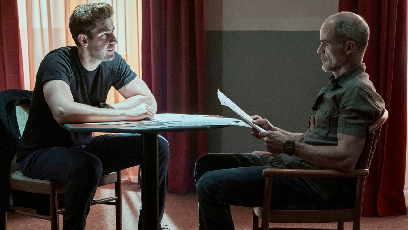 An image of two men sitting at a table in low light. One is holding several sheets of paper and they are looking at eachother.