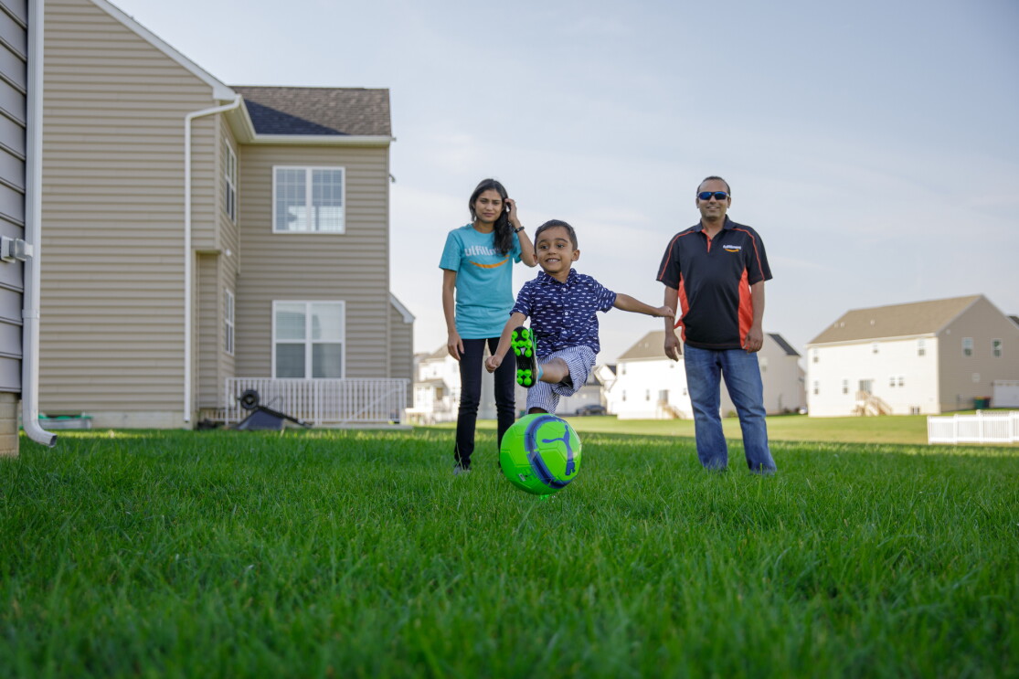 A young boy kicks a soccer ball on a lawn. He is flanked by a woman and a man, who watch as he plays. 