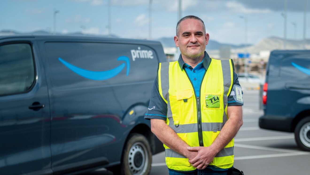Juan Antonio Carrasco, reconocido como Driver Hero. Está posando mirando a cámara, con las manos cruzadas delane y de fondo furgonetas de color gris con el logo, sonrisa de Amazon, en azul y la palabra Prime en blanco. Él está con las manos cruzadas delante, un polo azul de manga corta y una chaleco de color amarillo. Tiene el pelo corto negro y blanco. 