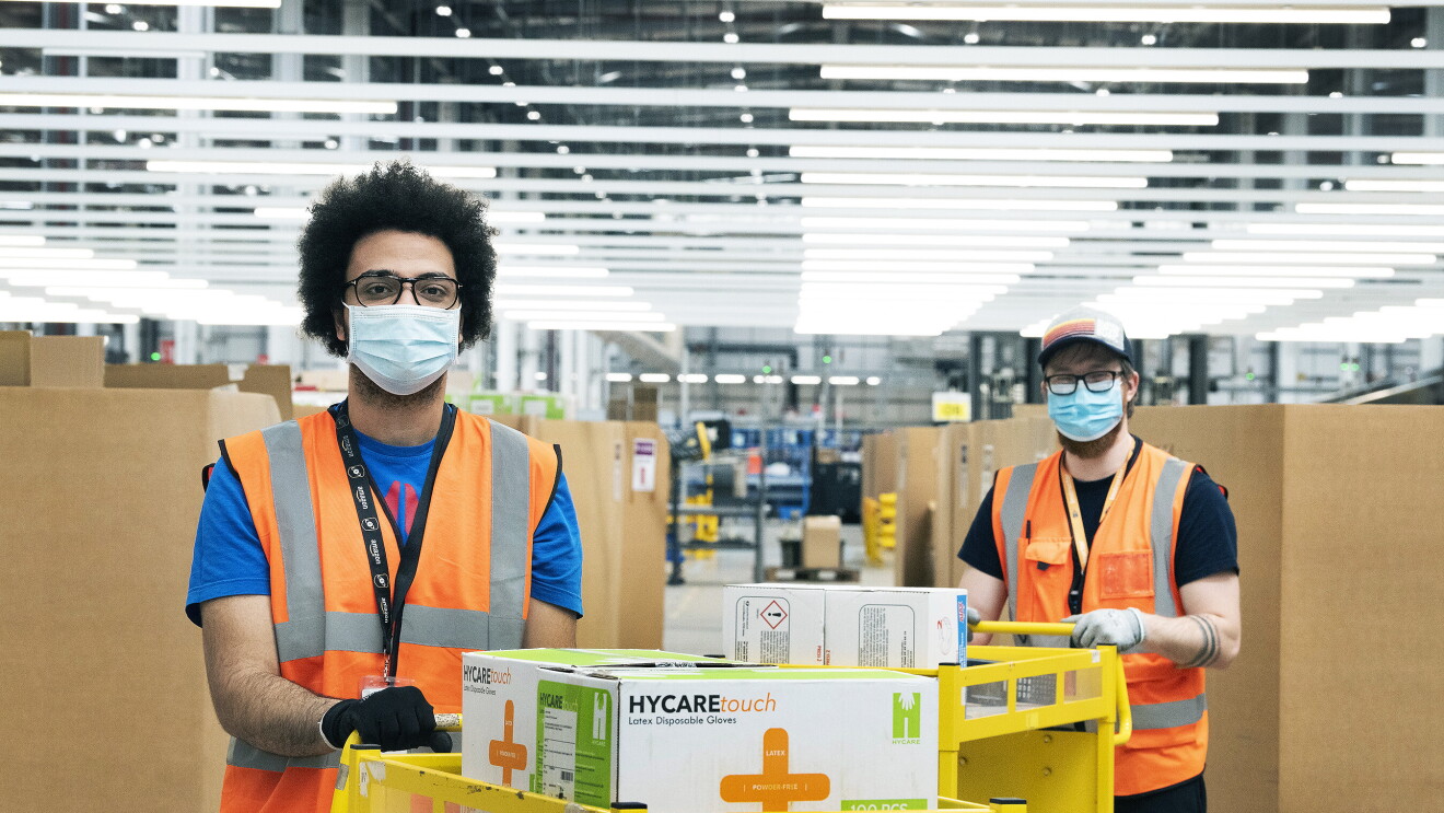 Amazon employees in a fulfilment centre packing medical supplies ready for the government into boxes. They are wearing hi-vis jackets and face masks. 