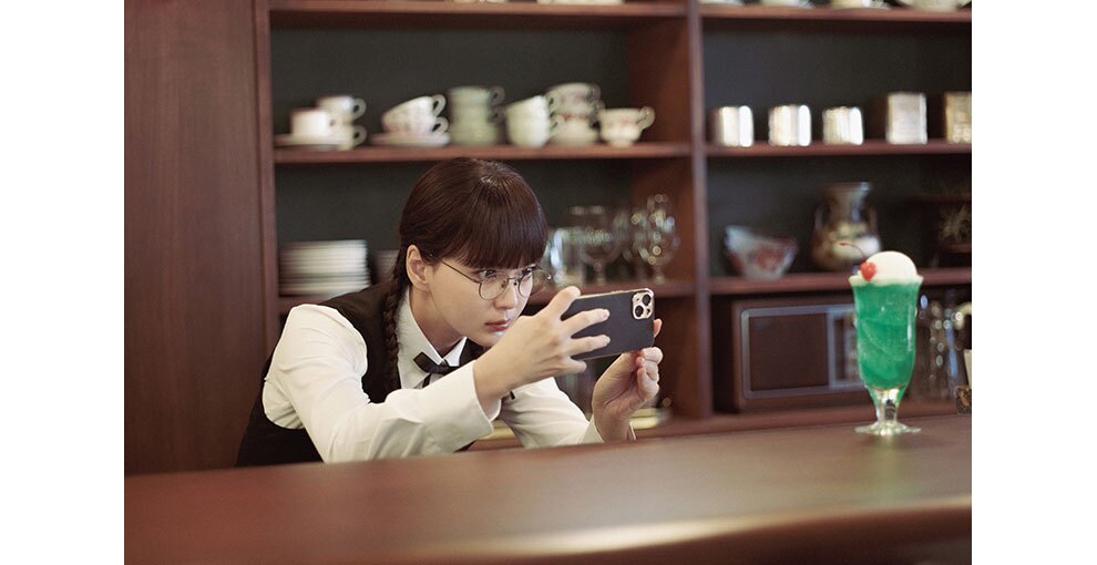 喫茶店のカウンターの中、ウェイトレスの服装をした若い女性が、カウンターの上のクリームソーダをスマートフォンで撮影している。