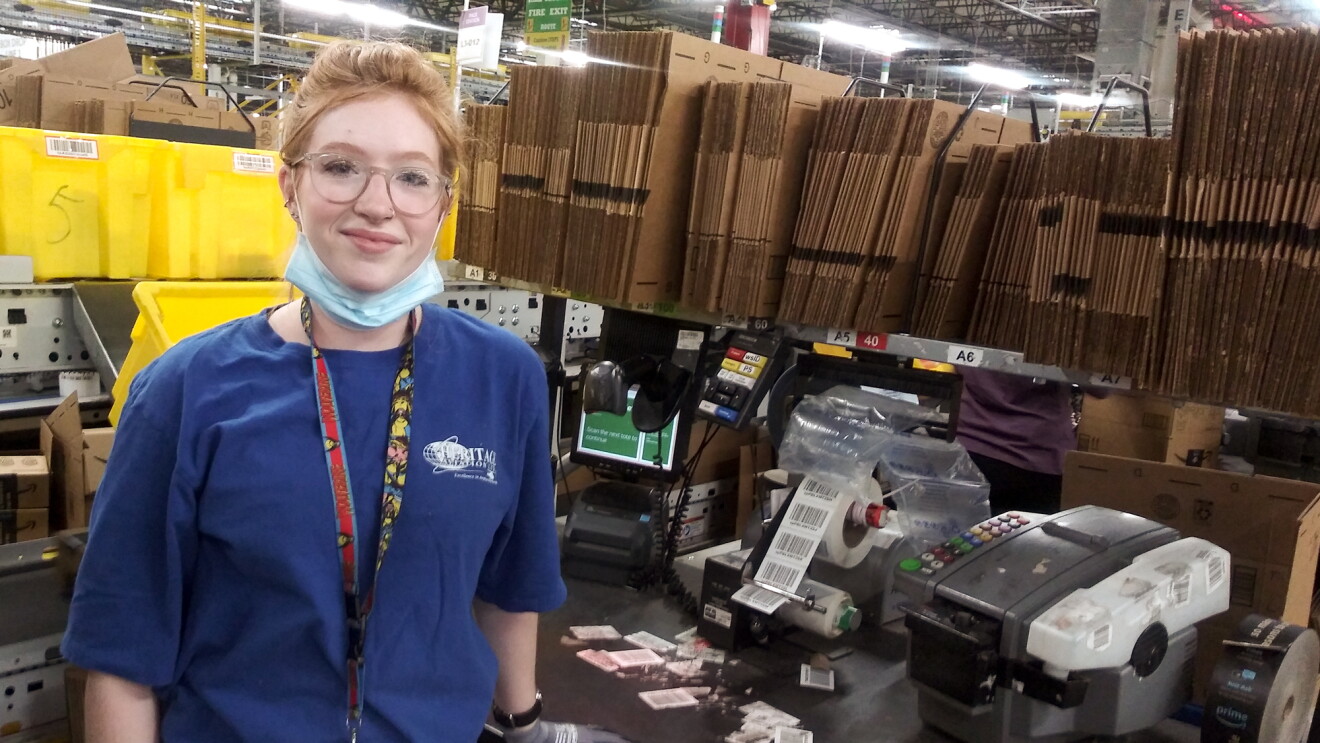 A woman stands at a workstation stocked with cardboard boxes and labeling machines. She has a protective mask on, ready to cover her mouth and nose.