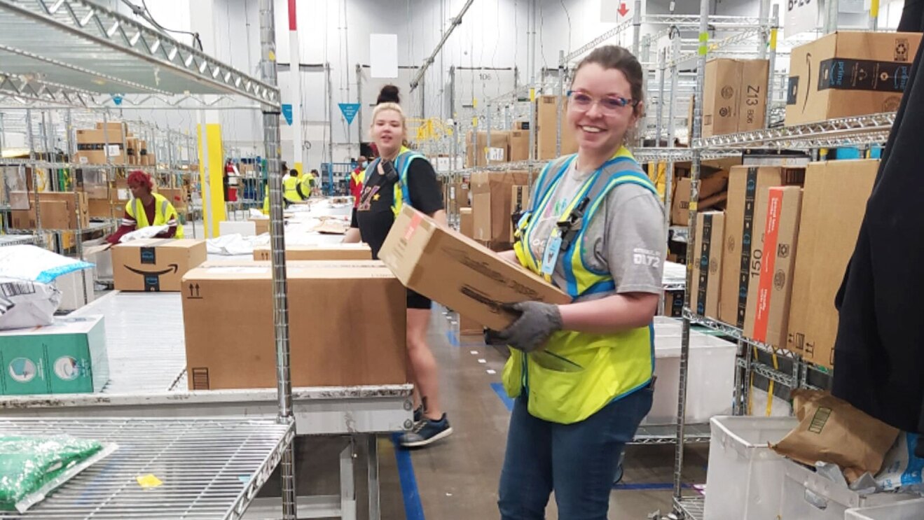 A woman wears a yellow safety vest and glasses and moves a box to a shelf in an Amazon fulfillment center.