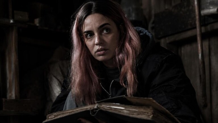Woman with pink hair holds a tattered book in a darkened room, looking out with a scared expression