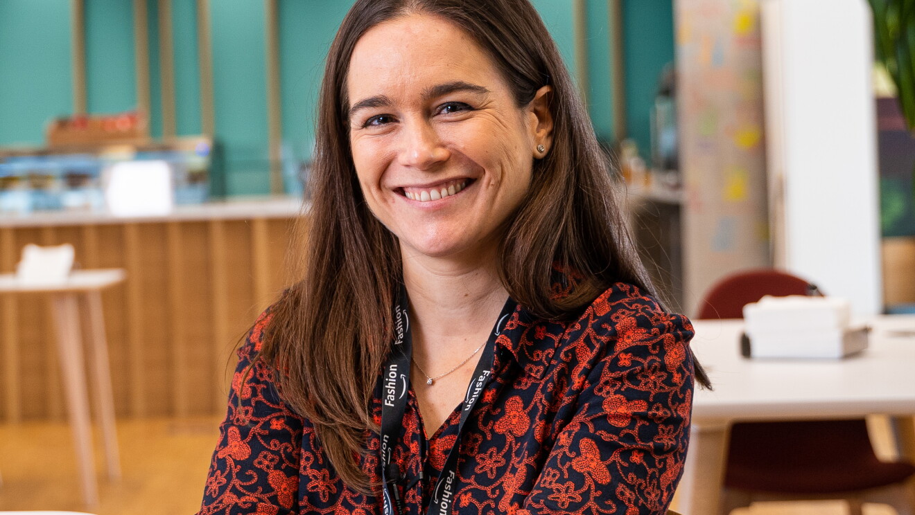 Lila Fernández es responsable de moda de los marketplace de Amazon en España e Italia.  De fondo está la cantina con mesas y los mostradores de comida. Lila mirando a cámara y sonriendo. Lleva un badge con una cadena de color negro. Una camisa azul con estampado de color rojo. Lleva pendientes y el pelo castaño, liso y largo. 