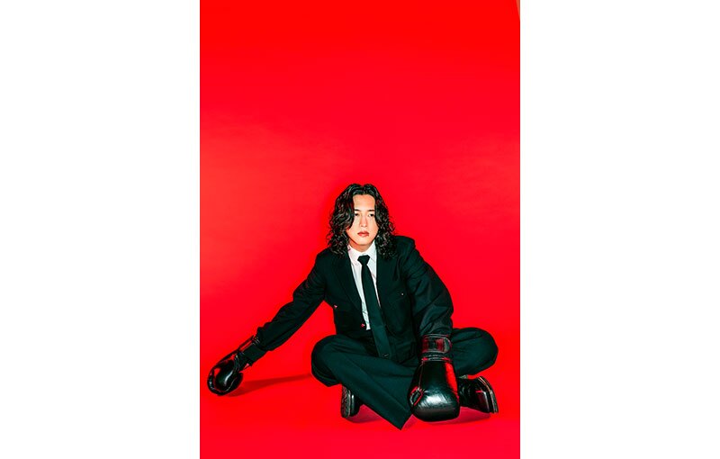 真っ赤な背景にあぐらをかいて座る黒スーツの男性。手にはボクシンググローブをはめている。