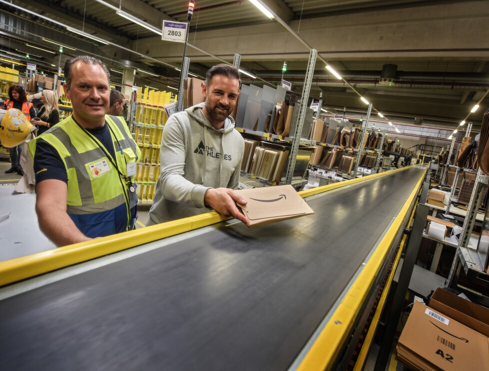 Christoph Metzelder legt ein frisch verpacktes Amazon Päckchen auf das Fließband im Logistikzentrum. Links von ihm steht Karsten Frost. Beide grinsen in die Kamera.