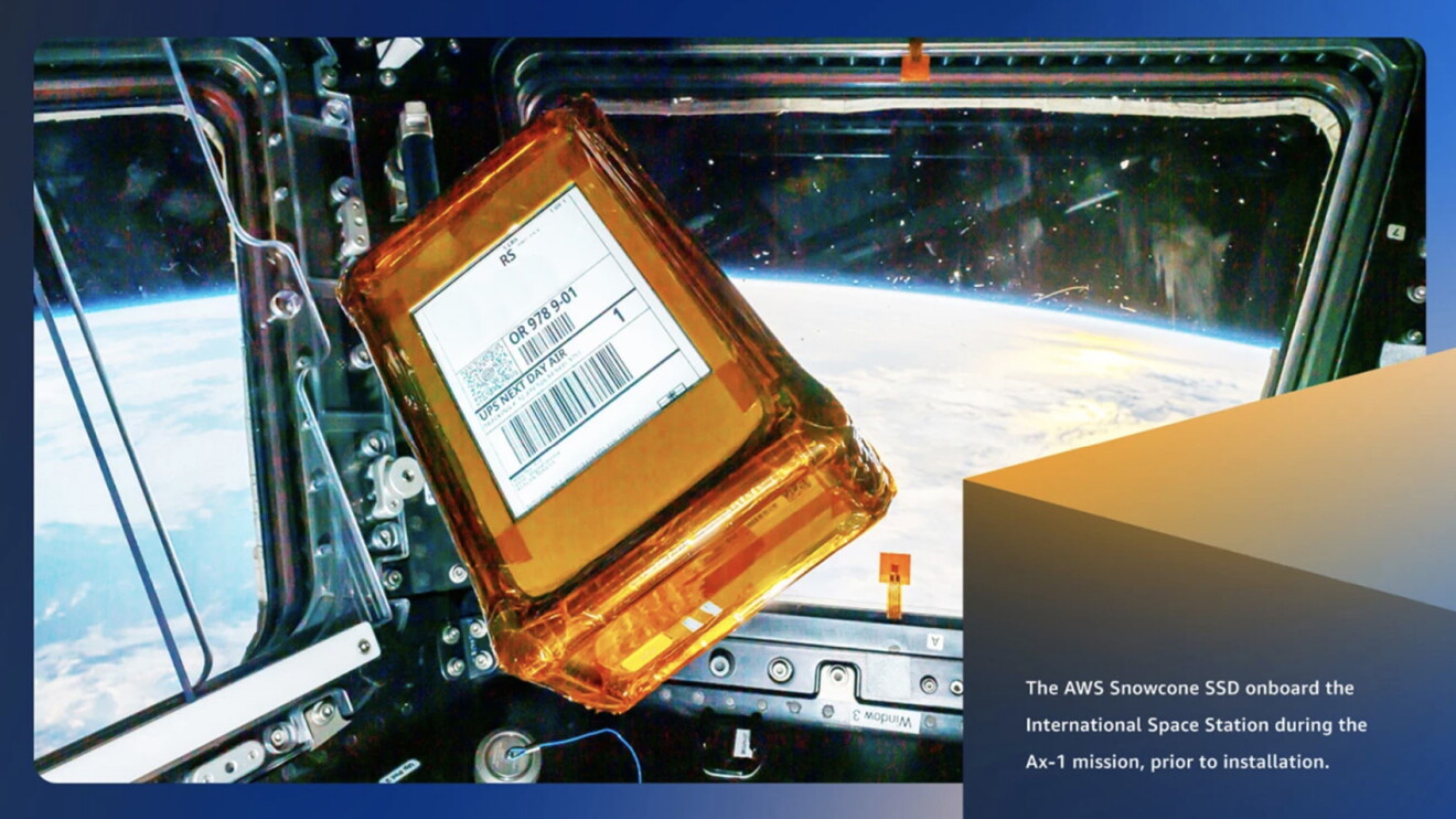 An image of an orange box floating in a space ship with windows overlooking earth and space. Text on the bottom right of the image says "The AWS snowcone SSD onboard the International Space Station during the Ax-1 mission, prior to installation." 