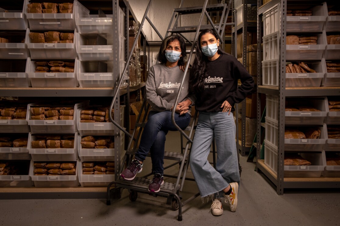 Two women in sweatshirts and jeans flanked by shelving in a warehouse space.
