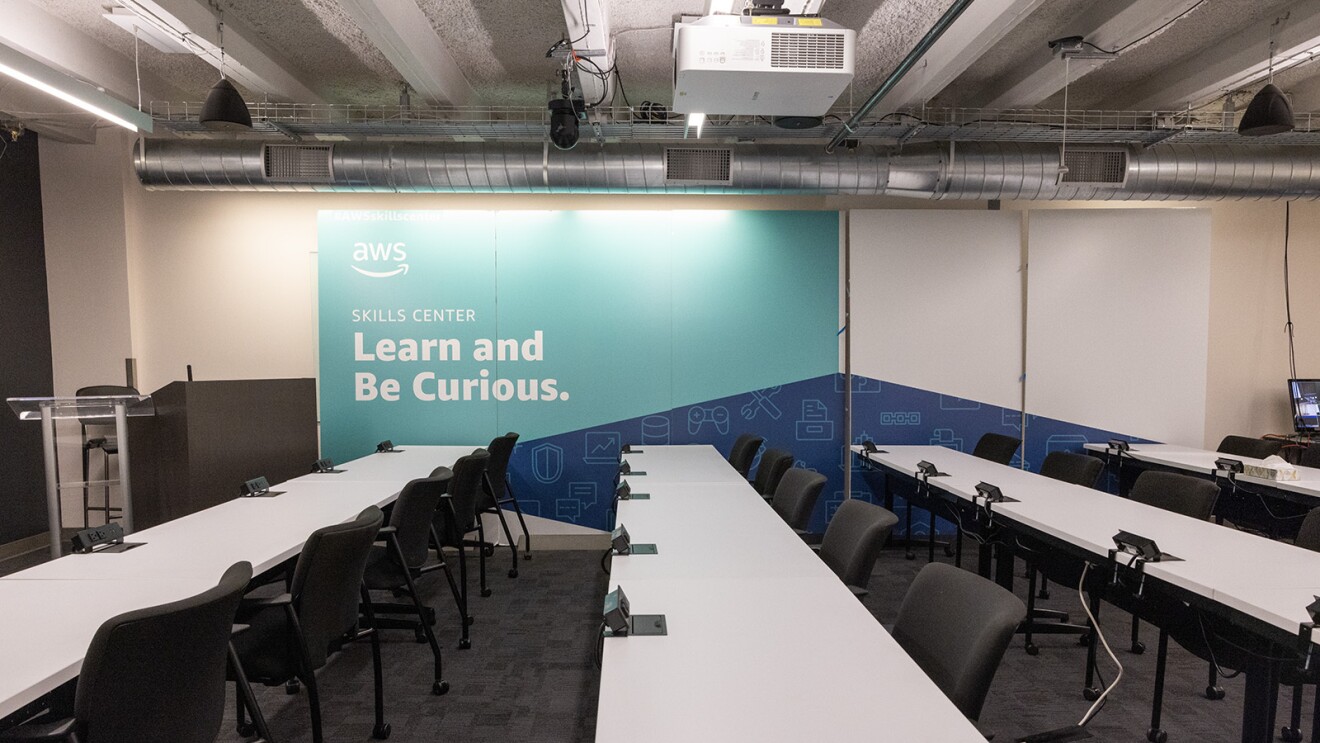 An image of the classroom inside the AWS Skills Center in Seattle. There are rows of tables with chairs seating at them and a wall that says "Skills Center: Learn and be curious" with the AWS logo