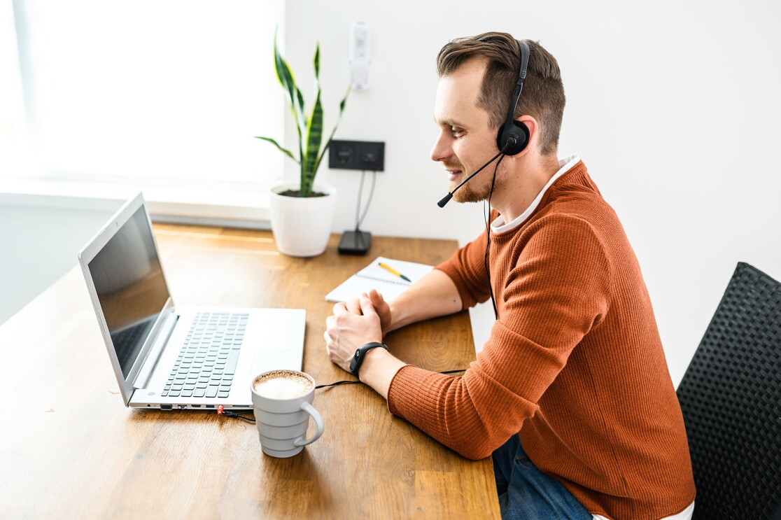 A man wears a headset and works on a laptop computer, with a notepad and coffee mug nearby. 