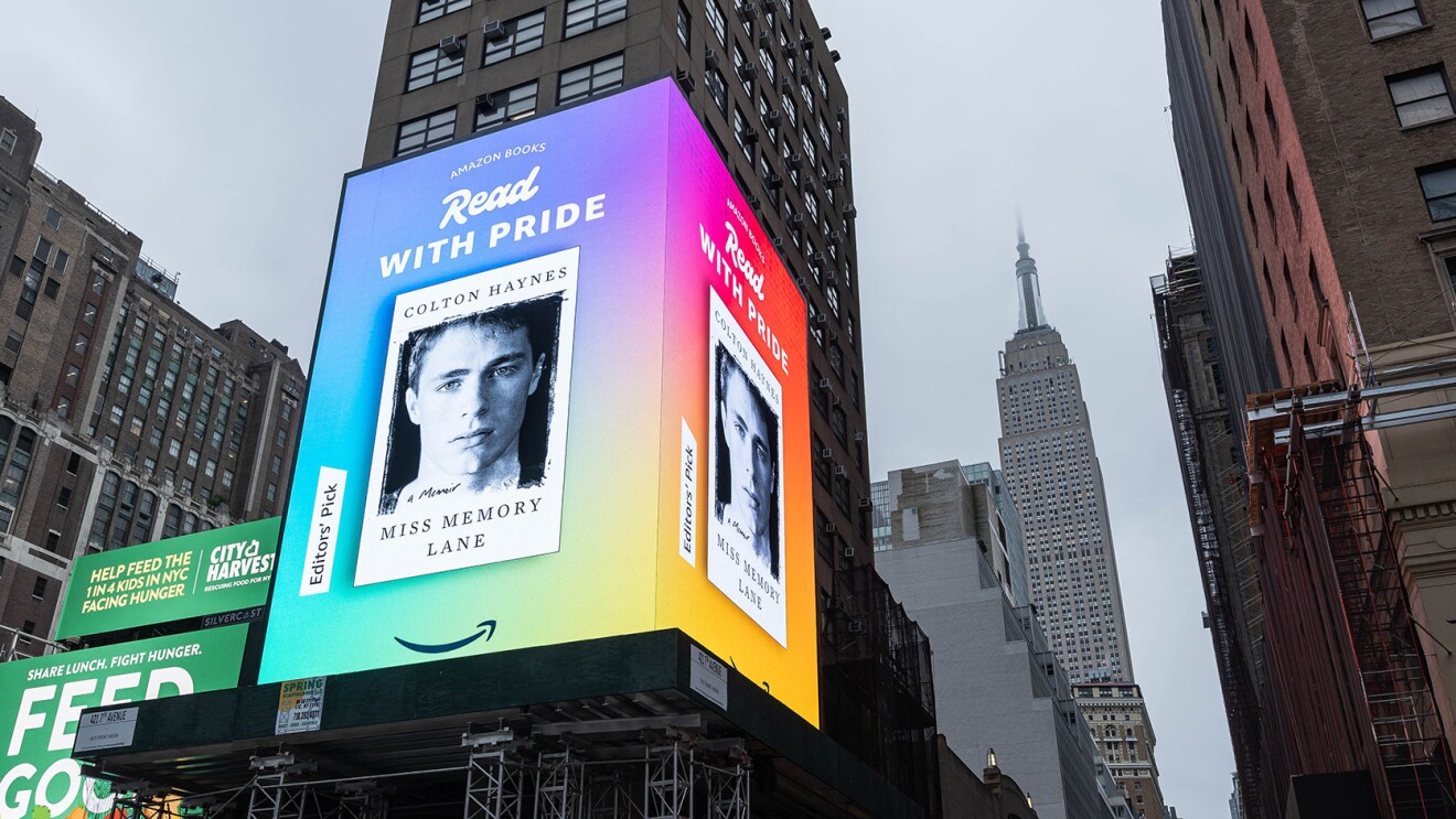 An image of a Times Square billboard with a book cover on it for "Miss Memory Lane." The text above the cover says "Read with Pride," and there is a tag on the side of the cover that says "Editor's pick." There is a rainbow-colored background behind the book and the Amazon smile logo below.