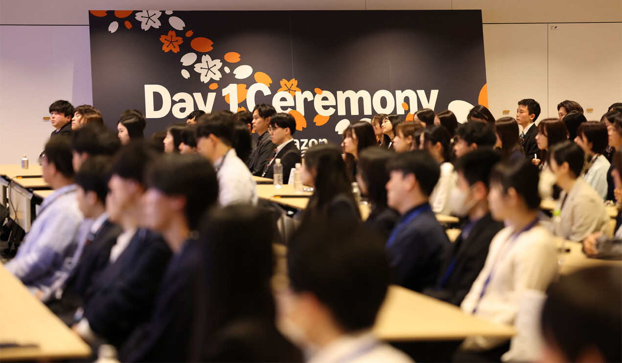 会議室の中で、黒い地にDay 1 ceremonyと白い文字で書かれた大きなパネルの前にあるたくさんの長テーブルの座席に座っている若い人たち。
