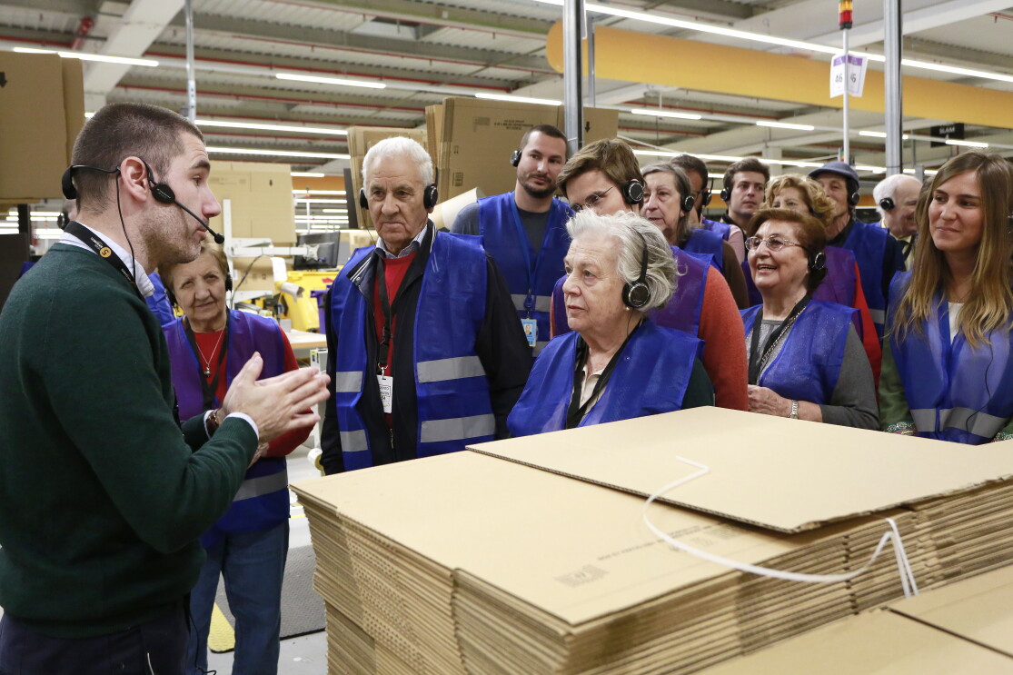 Detrás de unas cajas de Amazon se encuentra un grupo de personas mayores, abuelos, acompañados de su nietos, que escuchan atentamente las explicaciones del amazonian que les guía en su visita por el centro logístico.