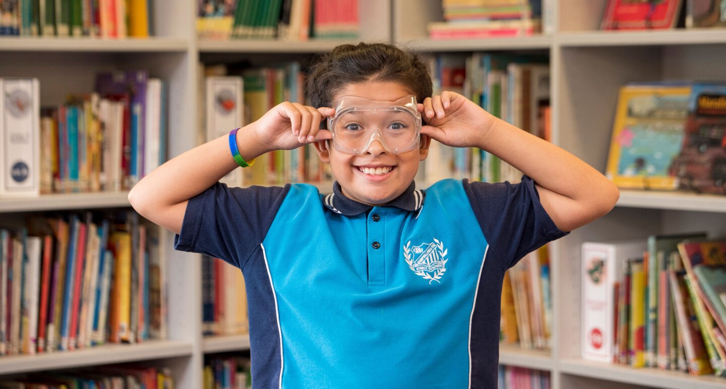 A young boy from Campbellfield Public School in Australia holds protective goggles to his face and gives the camera a goofy grin.