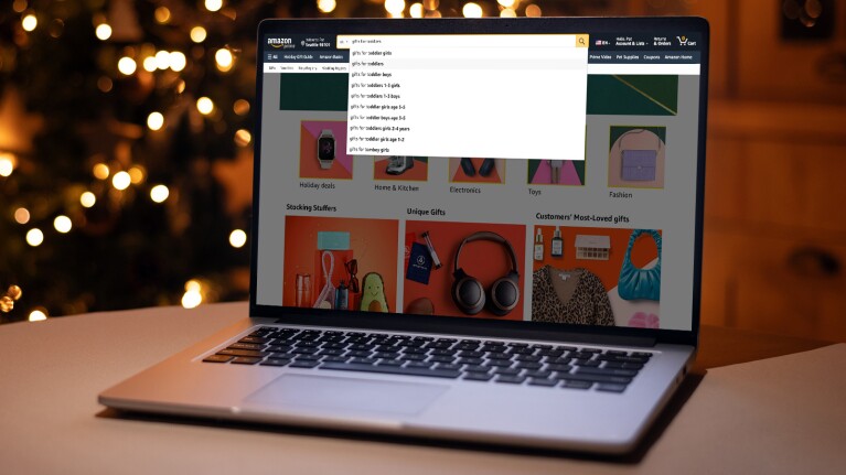 An image of a laptop sitting on a table. In the background you see a tree lit up with holiday lights. On the screen of the laptop you see someone searching on Amazon for gifts.