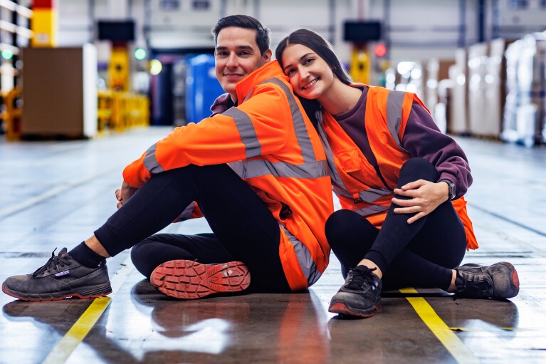 Fabiana y Rafael posando en el suelo de un centro logístico de Amazon. Los dos están posando, mirando de frente a cámara. Ella está de frente, los las piernas cruzadas, zapatos de seguridad negros, pantalón negro, camiseta morada y chaleco naranja. Lleva el suelo y es lacio y largo, Agarra con la mano derecha a Rafael. Él está de lado con las piernas cruzadas pero mirando a cámara. Lleva también zapatos de seguridad, pantalón negro, chaqueta que imita al chaleco de color naranja.