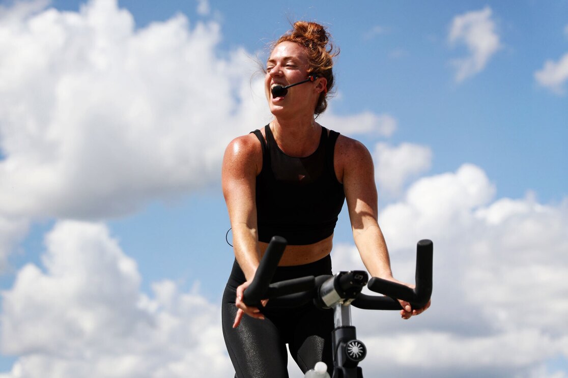 An image of a woman teaching a spin class outside. She is riding a stationary bike wearing a microphone and smiling while saying something to the class. There is a blue sky with light, white clouds behind her in the distance.