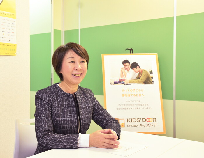ライトグリーンの壁を背に、白い椅子に座り白い机に両手を載せて話す女性。ネイビー地にチェック柄のスーツを着用。