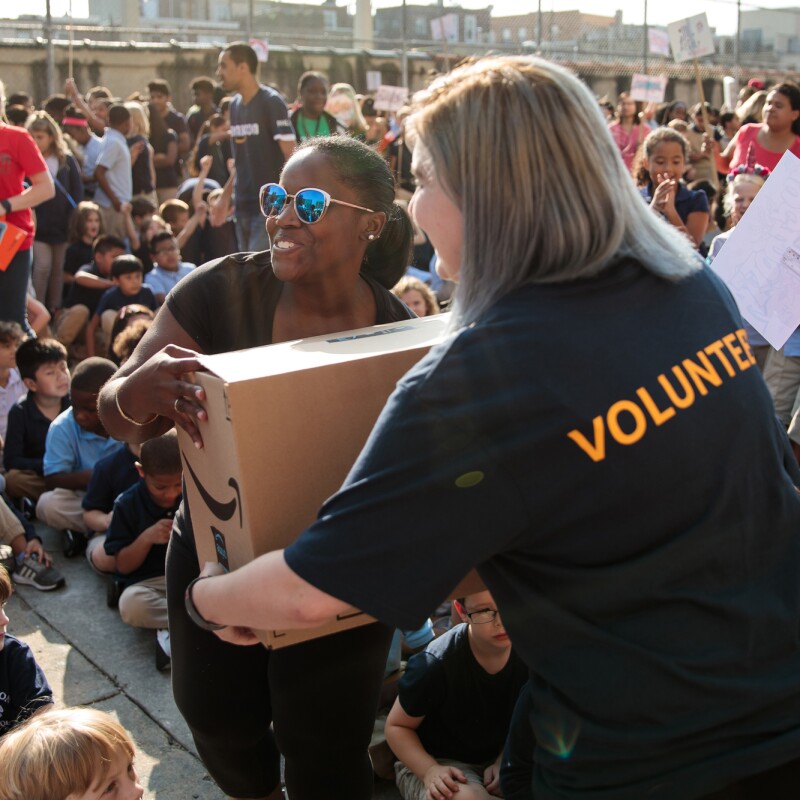 Two adult women carry a cardboard box marked with an Amazon logo. Seated children take up most of the image.