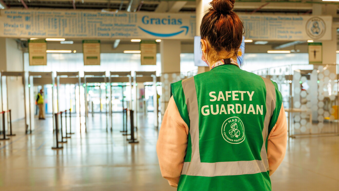 En la entrada de un centro logístico de Amazon aparece un Safety Guardian de espaldas. Es una mujer con una coleta recogida alta. La persona lleva mascarilla y un colar del que lleva el badge colgado. Tiene un chaleco de color de color verde con las letras blancas Safety Guardian MAD7.  