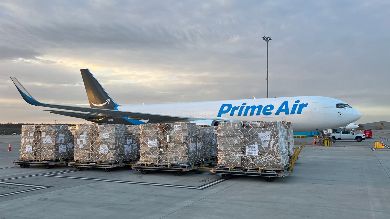 Pallets filled with boxes of humanitarian goods sit on the tarmac. An Amazon Air cargo plan is behind the pallets.