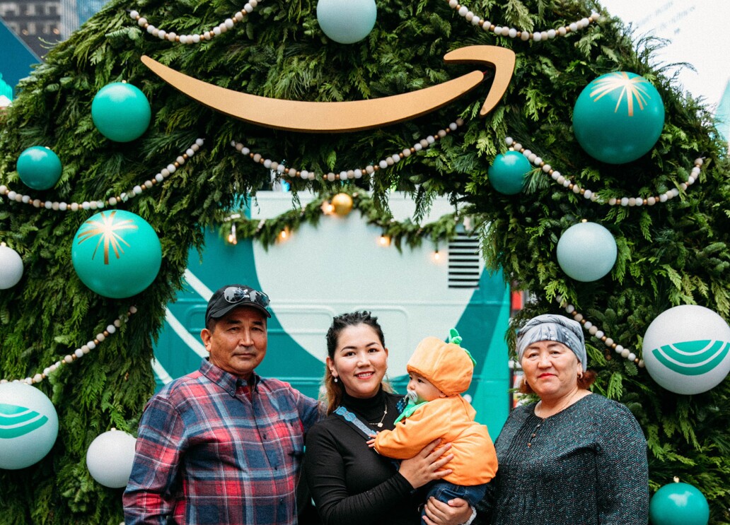 An image of a family smiling for a photo in front of a large wreath. There is a baby in the photo wearing an orange pumpkin costume.