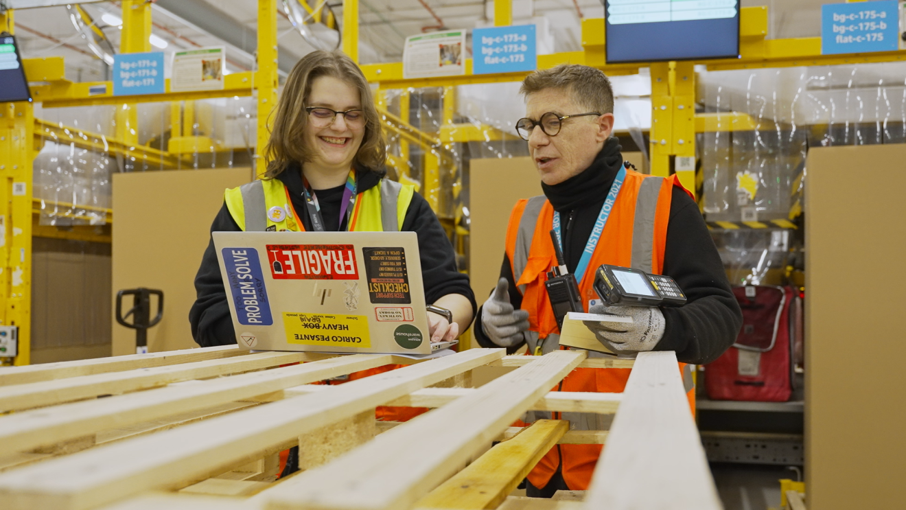 A young woman and a man with yellow and orange vests are looking at something on a laptop screen inside a fulfillment center