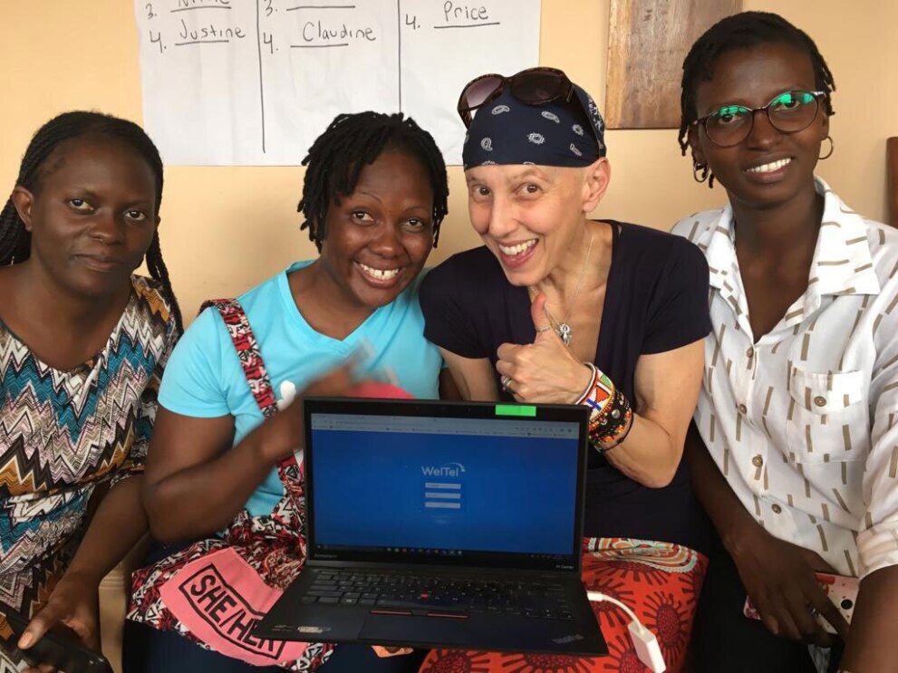 An image of four women smiling for a photo. There is a laptop in front of them and on the screen is the login screen for "WeTel"