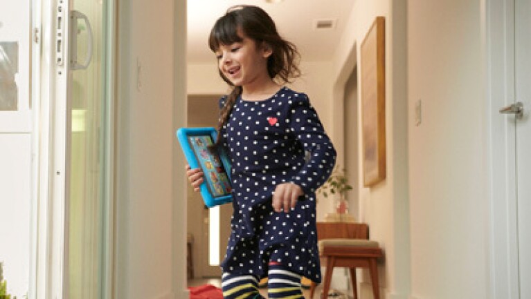 Young girl running through hallway holding an Amazon Fire tablet.