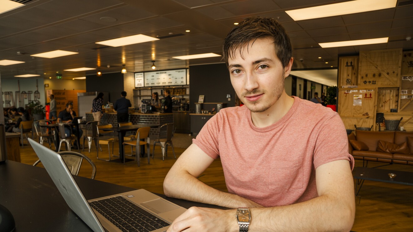 Jake Peters, 23, an Infrastructure Technician apprentice sitting in the UK Amazon corporate office cafeteria behind a laptop.